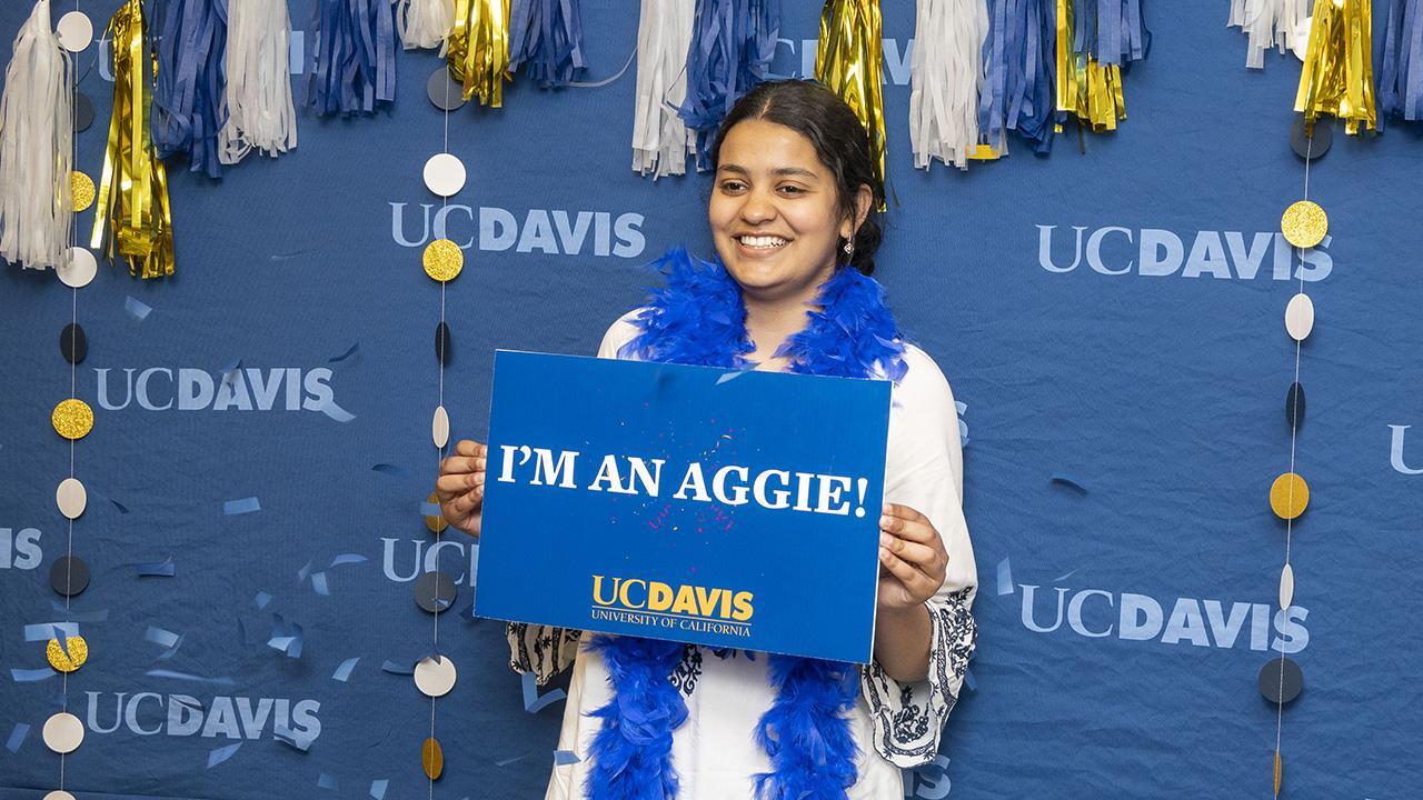 A person in a white tunic dress and blue boa, celebrates and poses for a picture infant of a celebratory UC Davis backdrop on Aggie Day 2024.
