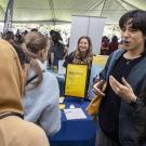 Two students one in a blue zip up sweatshirt and the other in a tan hoodie, talk among a crowd of people under a white tent at a resource fair on Aggie Day 2024.