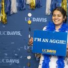 A person in a white tunic dress and blue boa, celebrates and poses for a picture infant of a celebratory UC Davis backdrop on Aggie Day 2024.